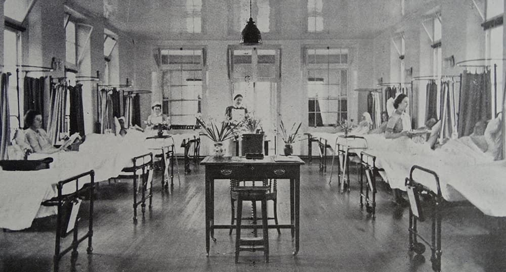 An NHS hospital ward from 1949 with two adjacent rows of beds and a desk in the middle. Nurses are tending to patients.